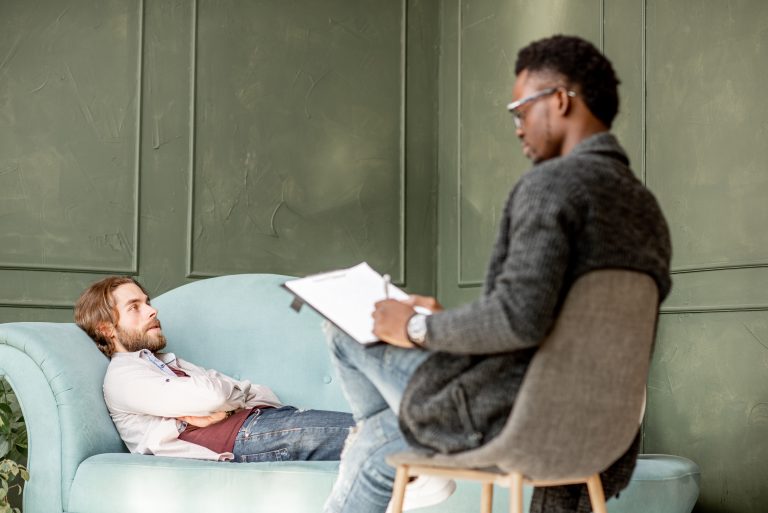 A male client reclines on a couch while a therapist takes notes, providing professional counseling support in a private and comfortable setting.