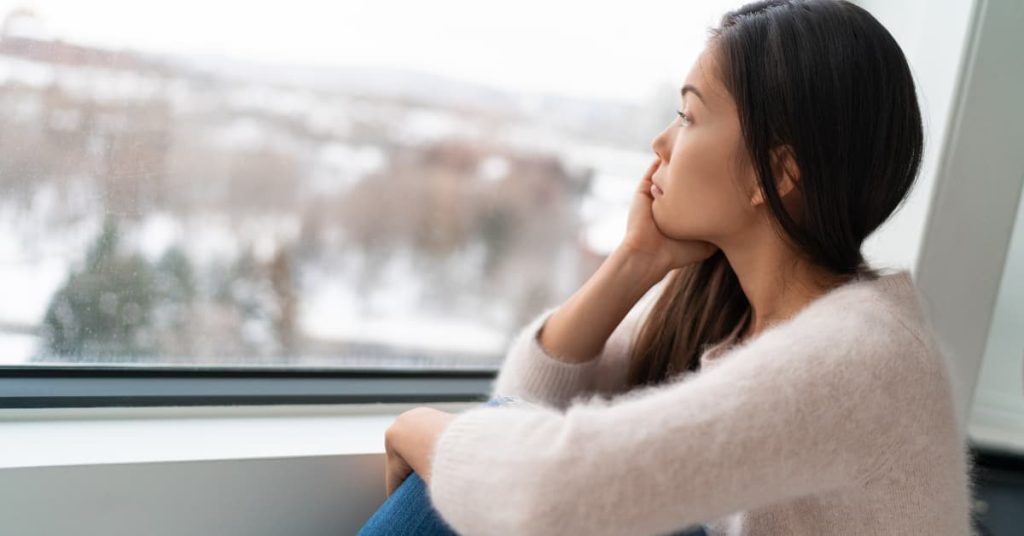 Thoughtful woman sitting by a window, staring outside at a blurred winter landscape, representing feelings of loneliness, sadness, or seasonal depression