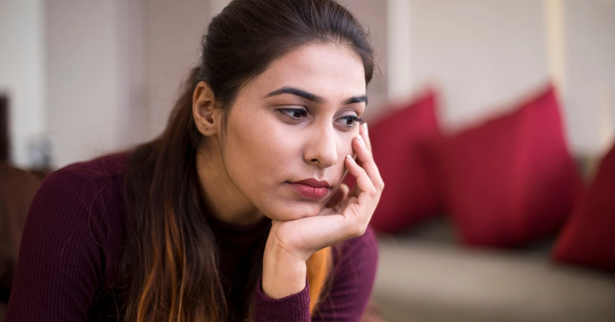 A young woman sitting indoors with a thoughtful, distressed expression, resting her chin on her hand—illustrating emotional struggle or maladaptive behavior.