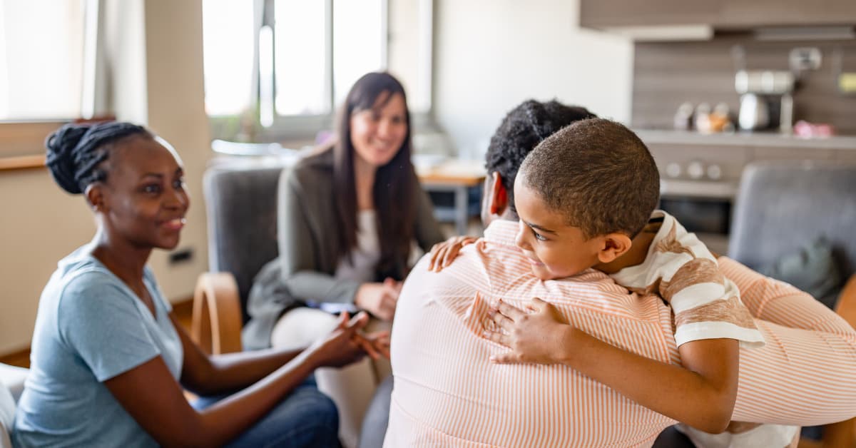 Smiling child hugging a supportive adult during a counseling session, with mental health professionals and caregivers present in a warm, home-like setting.